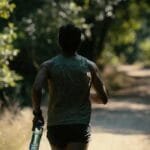 Indian runner holding electrolyte hydration bottle while running on a scenic trail during hot weather training in India, back view with green trees and dirt path
