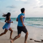 A young man and woman running together barefoot on a scenic Kerala beach at sunset, with turquoise ocean waves, palm trees in the background, and golden sand underfoot – perfect for marathon training in South India