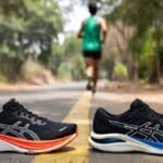 Hoka Clifton 10 and Asics Gel-Nimbus 27 max-cushion running shoes placed on an Indian dirt road, with a runner jogging in the background wearing green tank top during hot weather marathon training in Greater Noida 2026