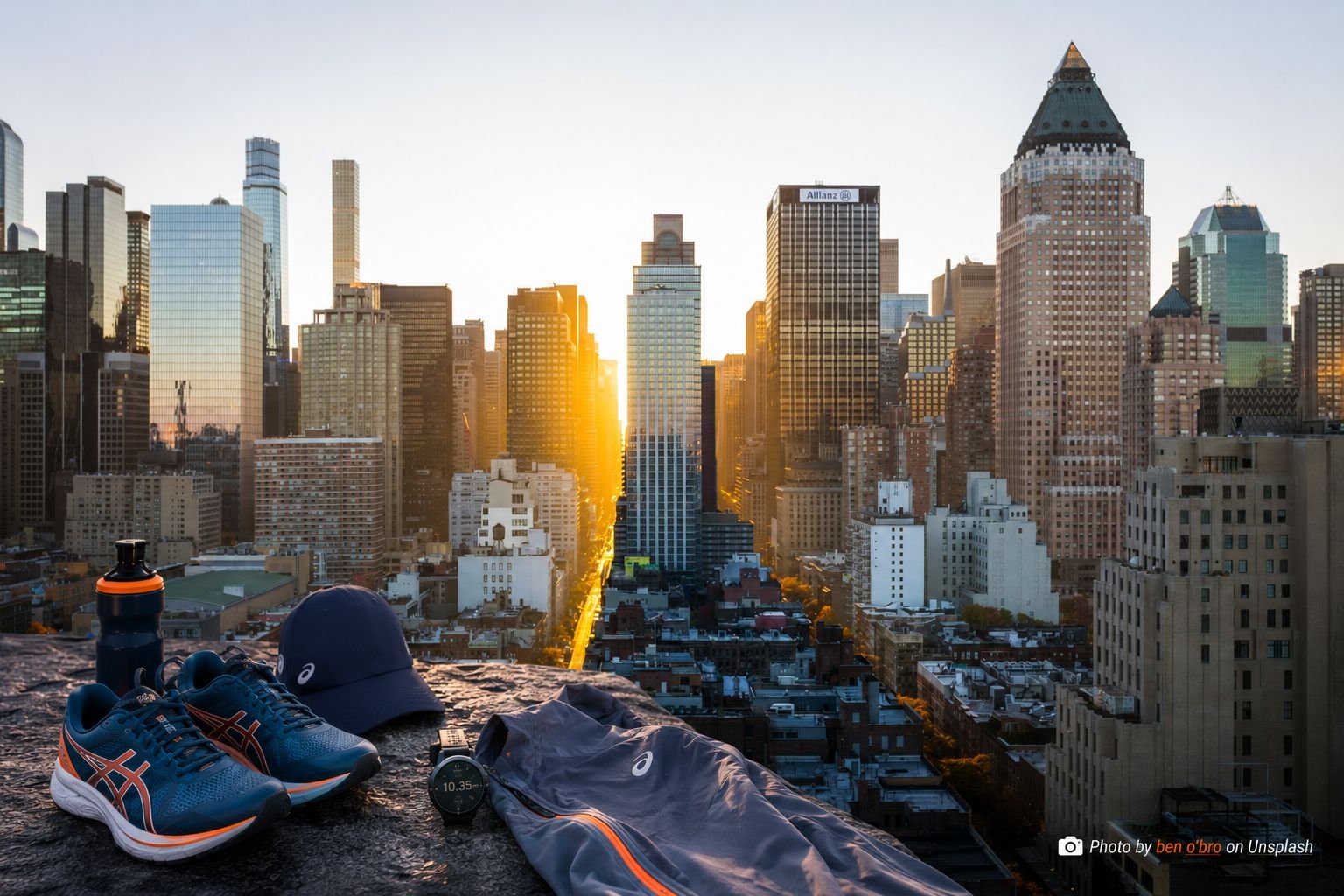 Marathon training gear including ASICS running shoes, GPS watch and hydration bottle laid out against the New York City skyline at sunrise