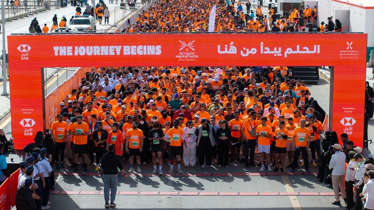 Thousands of runners in orange shirts at the Riyadh Marathon start line under a banner reading "The Journey Begins" at the mass race start in Saudi Arabia
