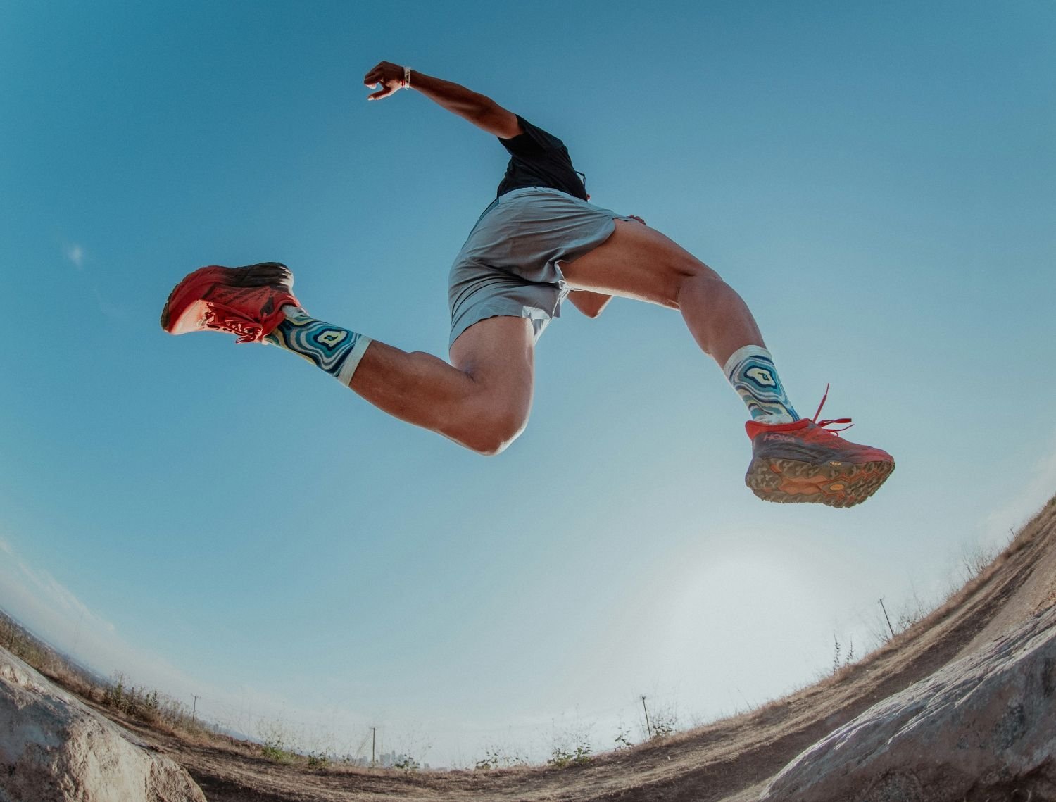 Trail runner leaping mid-air over dirt terrain wearing cushioned red Hoka trail running shoes and patterned socks.