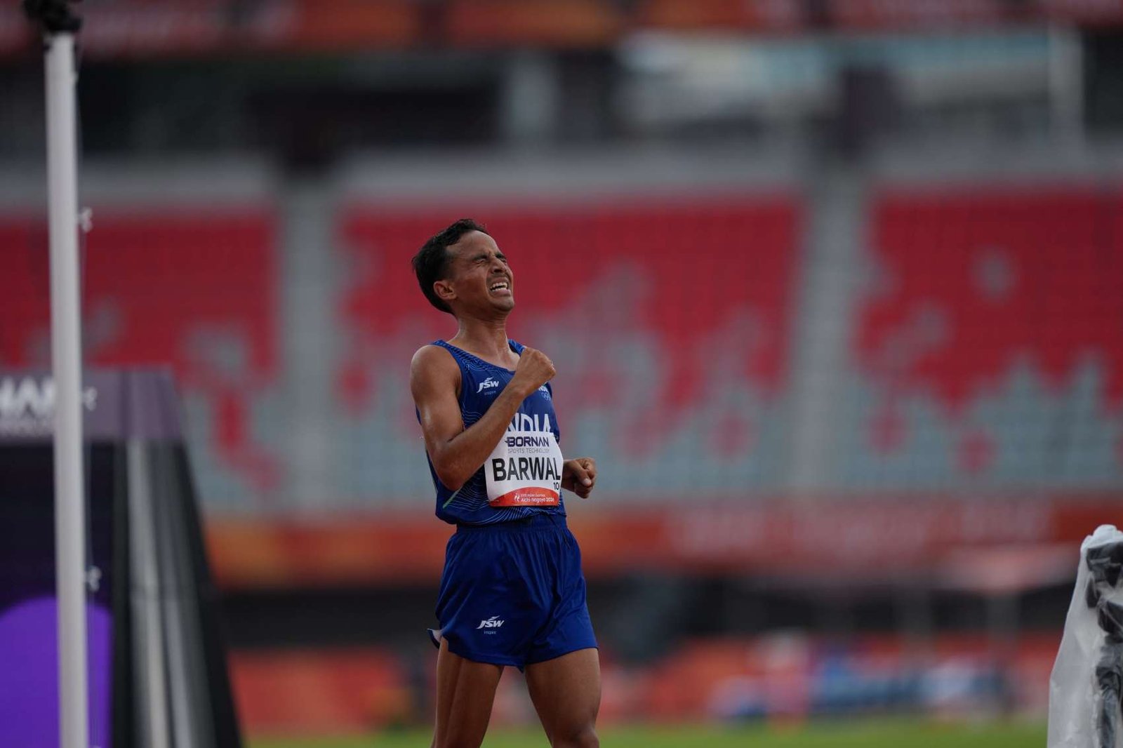 Sawan Barwal celebrates at the finish line of the men's marathon at Asian Games 2026 in Nagoya, wearing India's blue JSW kit and his race bib, fist clenched as he clinches silver with a new national record of 2:11:37
