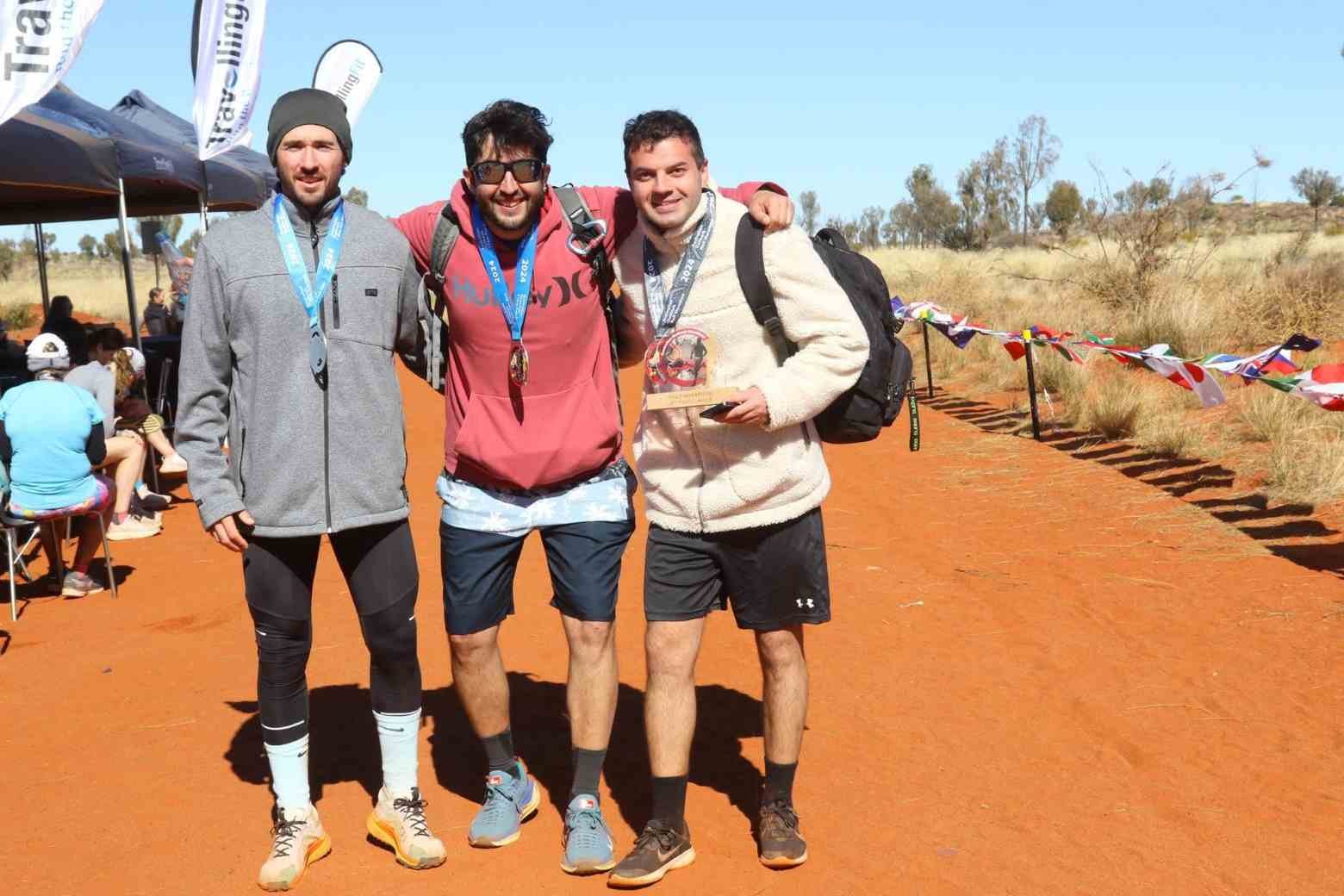 Three finishers celebrate with medals and a trophy at the Australian Outback Marathon finish line on the red earth at Uluru