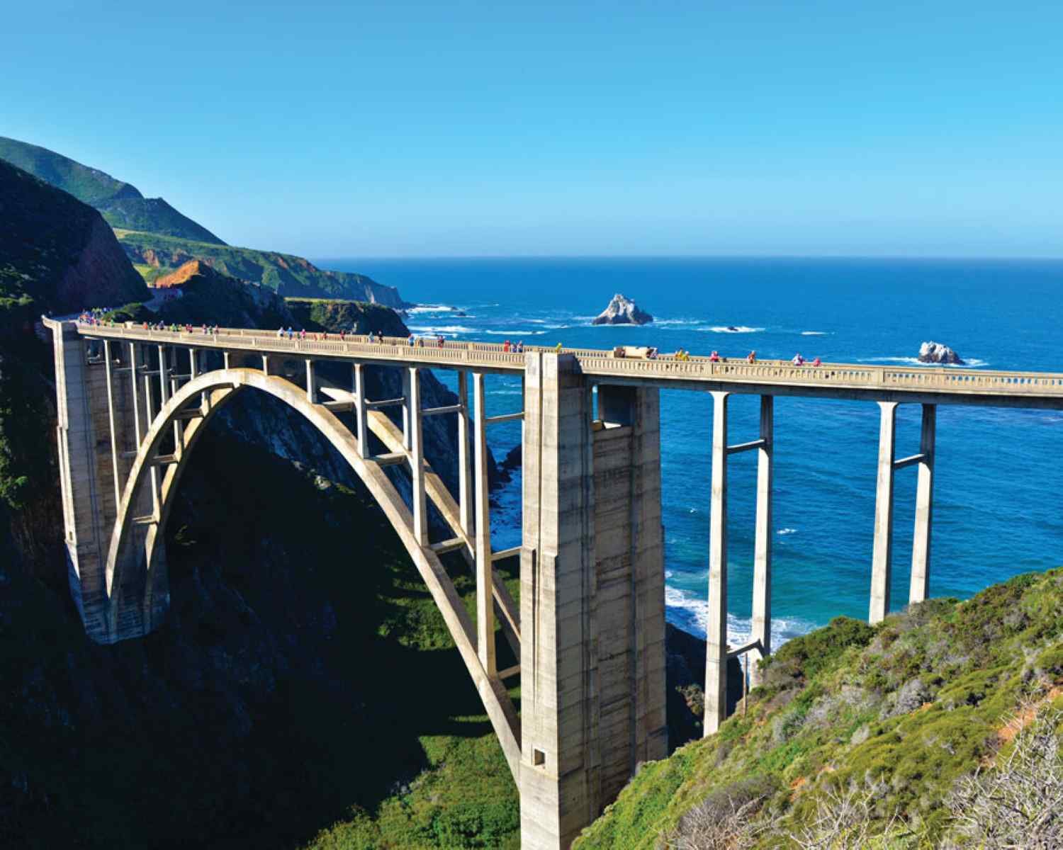 Runners crossing Bixby Bridge during the Big Sur International Marathon, with the Pacific Ocean and coastal cliffs in the background