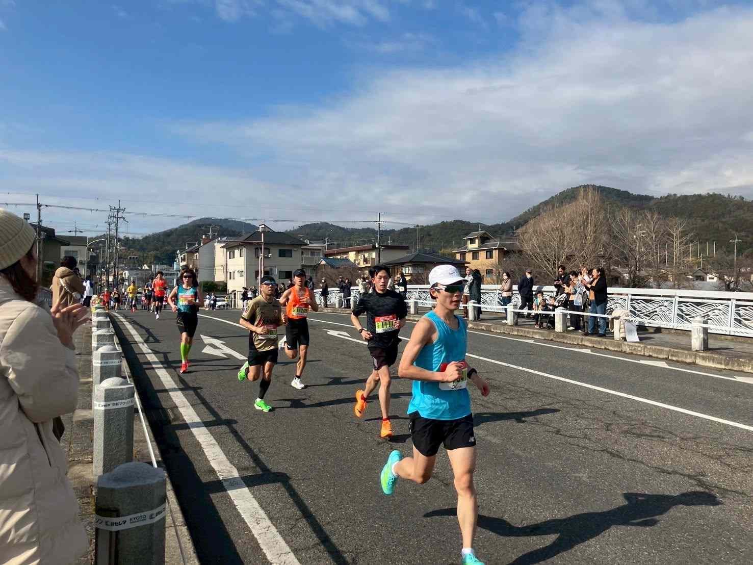 Runners crossing a bridge on the Kyoto Marathon course with Arashiyama hills in the background