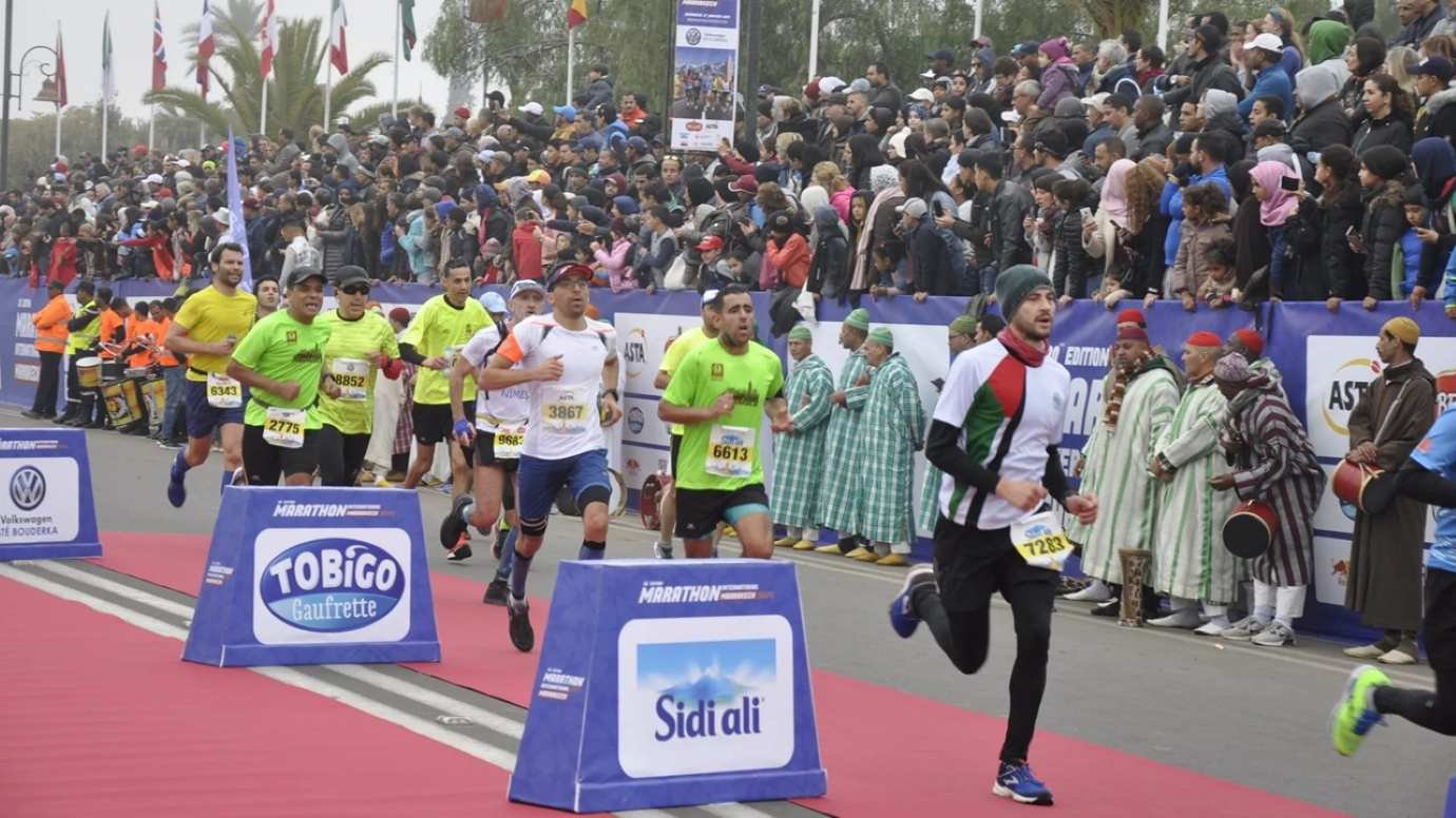 Runners crossing the finish line at the Marrakech International Marathon with a packed crowd and traditional Moroccan musicians lining the route