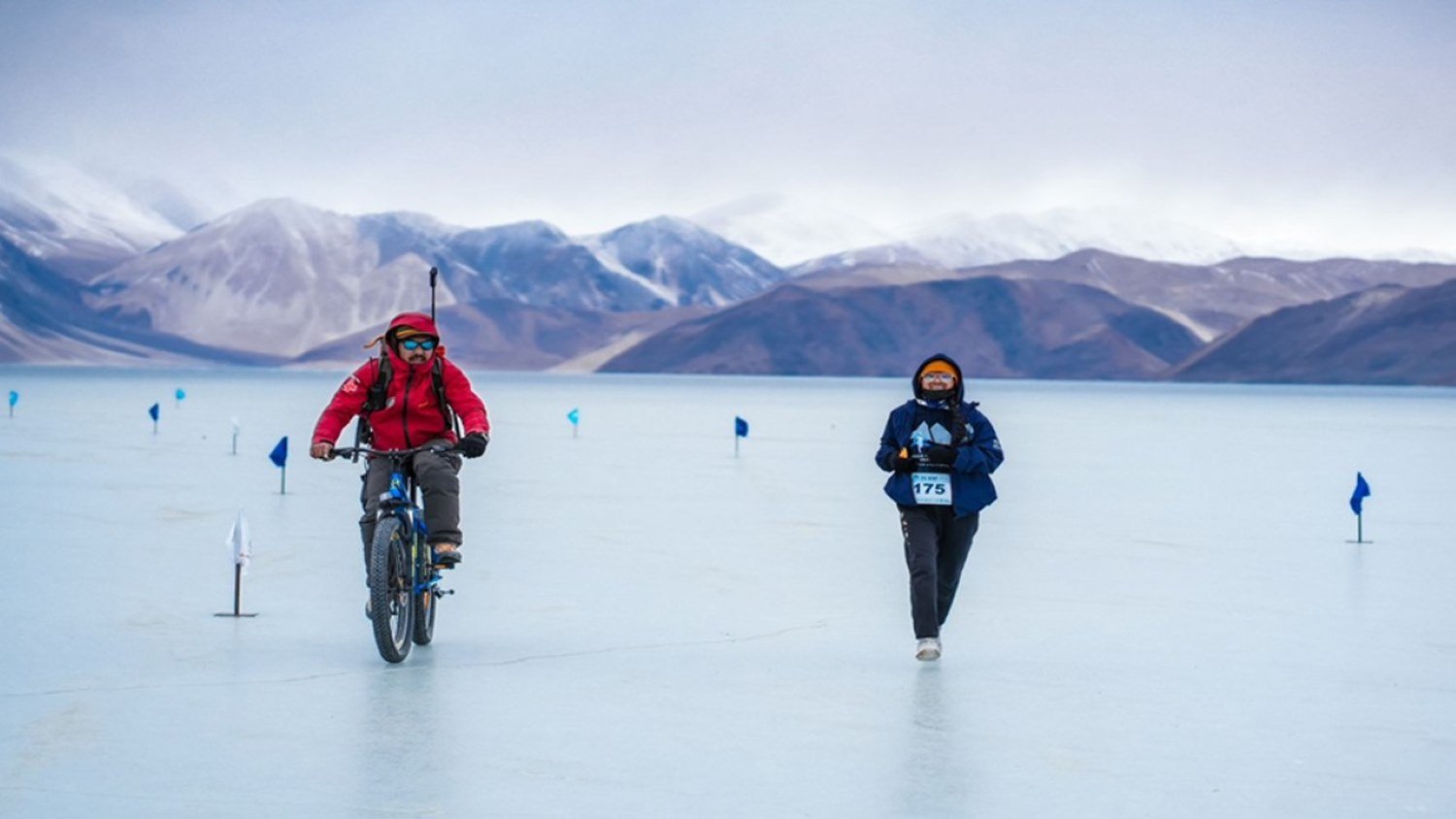 A runner wearing BIB 175 runs on the frozen surface of Pangong Tso escorted by a cyclist on the Pangong Frozen Lake Marathon course, with snow-capped Himalayan peaks in the background